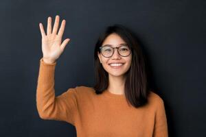 Young woman with glasses smiling and waving against a dark background in an indoor setting photo