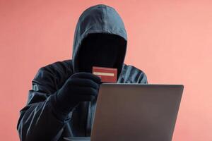 Anonymous individual engaged in cyber activity with credit card in front of laptop on a solid color background photo