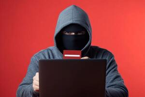 Cybercriminal wearing a hoodie engages in identity theft while using a laptop against a red background photo