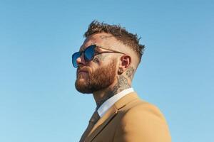 Man in a beige suit and sunglasses displays confident style against a clear blue sky photo
