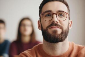 Young man with glasses attentively listening in a classroom setting during a lecture with classmates in the background photo