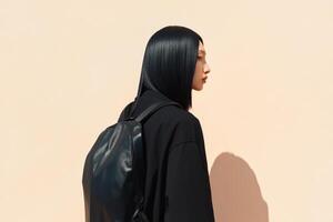 Young woman with sleek black hair and backpack poses against a peach wall in bright daylight photo
