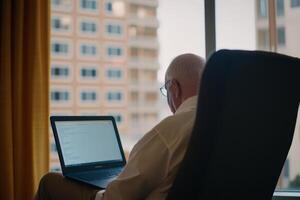 Elderly man working on laptop in cozy room with city view during the day photo