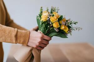 Person holds a bouquet of yellow roses wrapped in brown paper in a bright indoor setting during daytime photo