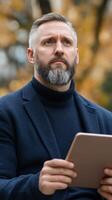 Man with a beard holding a tablet while deep in thought at a park during autumn photo