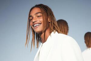 Young man with dreadlocks wearing a stylish white outfit smiles confidently against a clear sky backdrop in a modern setting photo