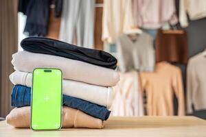 Stack of clothing prepared for resale with a smartphone displaying green screen for mockup use. Mobile apps simplify second-hand fashion, sustainable shopping, and conscious clothing redistribution. photo