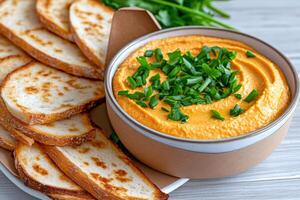 Bowl of red lentil hummus garnished with chopped chives next to toasted slices of baguette bread photo