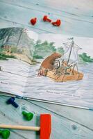 Ukraine, Lviv - March 04, 2025. Open book of fairy tales with drawn beavers sailing in a boat lies on a wooden table next to a toy hammer and bolts photo