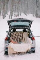 Car with an open trunk with a blanket, thermos, pillows and mugs stands in a snowy forest photo