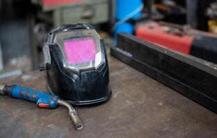 Close-up on the welding helmet, next to the left the welding gun. On the right two welded metal bars. Conceptual image of work, metalworking, craftsmanship. Copy space. photo