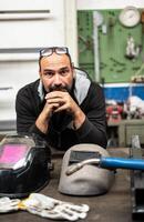 Nice portrait of a Caucasian man looking into camera. On his bench are his welder's work tools, two protective helmets and the welding gun. Vertical format. photo