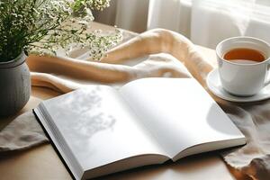Empty book for mockup with blank pages beside a cup of tea and a vase of flowers on a wooden table in a bright, cozy indoor setting during the afternoon photo