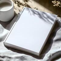 Blank book for mockup resting on soft fabric beside a cup of milk illuminated by natural light in a cozy indoor setting photo