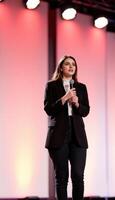 Confident speaker addresses audience with microphone against radiant backdrop on stage. Formal attire. photo