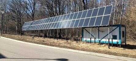 A solar panel array stands next to a small utility building in a wooded area. Sunlight reflects off the panels, emphasizing the commitment to renewable energy photo