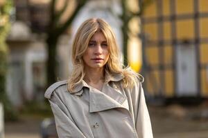 Elegant woman in light trench coat poses confidently on urban street with autumn vibes photo
