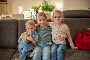 portrait of three children at home. brother and sisters sit on the couch hugging and smiling photo