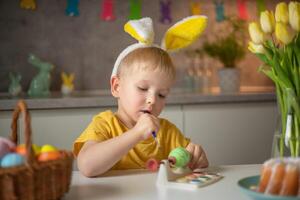 A little boy wearing bunny ears prepares painted Easter eggs for Easter while sitting at the kitchen table. Happy easter photo