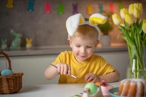 A little boy wearing bunny ears prepares painted Easter eggs for Easter while sitting at the kitchen table. Happy easter photo