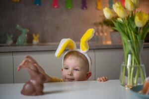 A cute little boy wearing bunny ears on Easter day wants to eat a chocolate easter bunny. A child plays egg hunt for Easter. Charming child celebrates Easter at home photo