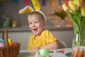 Emotional portrait of a cheerful little boy wearing bunny ears on Easter day who laughs merrily, plays with colorful Easter eggs sitting at a table in the kitchen photo