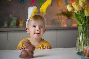 A cute little boy wearing bunny ears on Easter day wants to eat a chocolate easter bunny. A child plays egg hunt for Easter. Charming child celebrates Easter at home photo