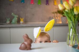 A cute little boy wearing bunny ears on Easter day wants to eat a chocolate easter bunny. A child plays egg hunt for Easter. Charming child celebrates Easter at home photo
