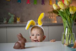 A cute little boy wearing bunny ears on Easter day wants to eat a chocolate easter bunny. A child plays egg hunt for Easter. Charming child celebrates Easter at home photo