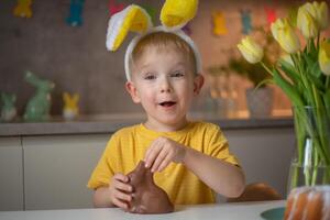 A cute little boy wearing bunny ears on Easter day is eating a chocolate Easter bunny. A child plays egg hunt for Easter. Charming child celebrates Easter at home photo