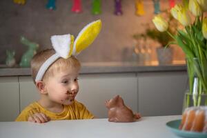A cute little boy wearing bunny ears on Easter day is eating a chocolate Easter bunny. A child plays egg hunt for Easter. Charming child celebrates Easter at home photo
