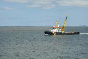 A utility vessel navigates smooth waters near a coastal region on a clear day, showcasing efficient marine operation and maintenance activities photo