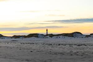 Bright lighthouse stands tall over sandy beach at sunset, casting a serene glow on the coastal landscape photo