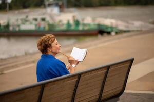 Woman in a blue shirt reading while seated on a bench by the river photo