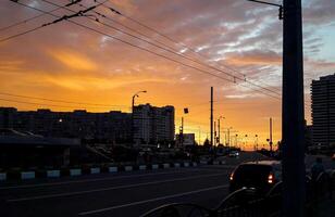 Vibrant urban sunset scene with silhouettes of buildings, cars, power lines, and dramatic orange sky. Captures the evening atmosphere of a modern city with street life and infrastructure. photo