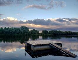 Peaceful rural river scene with wooden raft, sunset sky and forest reflected in calm water. Natural landscape with soft colors and atmospheric clouds. photo
