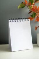 Blank notepad with spiral binding placed on a table next to a potted plant showcasing autumn-colored leaves photo
