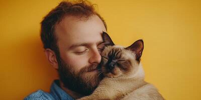 Man enjoys cuddle time with his Siamese cat against a vibrant yellow backdrop in a warm indoor setting photo