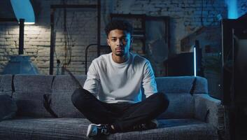 Young man sits cross-legged on a couch in a dimly lit room with blue lighting and an industrial background photo