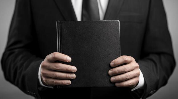 Businessman holding a black notebook in a formal suit while standing against a neutral background photo