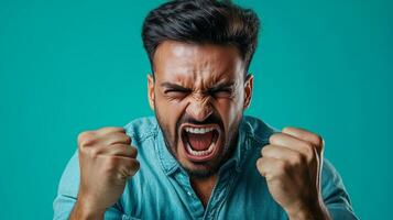 Man expressing intense emotions with clenched fists and a fierce shout against a solid turquoise background photo