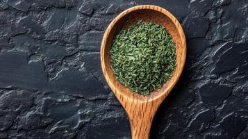 Dried green herb in wooden spoon on textured dark surface for culinary use photo