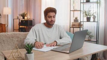 Young Indian man student attending online class through laptop while taking notes at table at home video