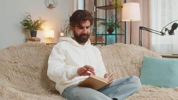 Indian man reading interesting book, turning pages smiling enjoying literature sitting on sofa video