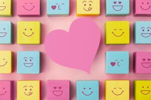 Love and Human Relationship Concepts. Happy Community. Pastel Pink Heart Shape surrounded by Group of Smiling Emoticon Diversity Face on Cube. Table Top View photo