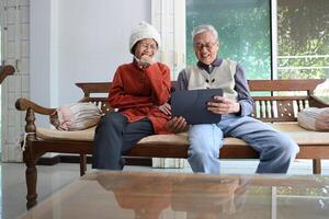 Senior asian couple are laughing and sitting on the sofa while watching funny stand up comedy show from streaming digital tablet during winter for mental health and happiness in marriage photo