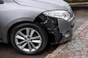 Front of a silver car with a damaged bumper and exposed inner structure after an accident, parked near a curb on a wet road photo