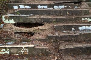 Old crumbling concrete stairs with large holes, cracks, and peeling paint, surrounded by dirt and debris, showing decay photo