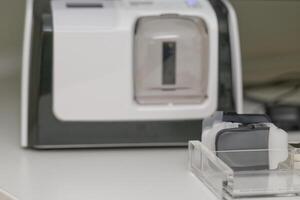 A closeup view of a medical device xray scanner with a transparent chamber and various attachments. The background features cables and other equipment, suggesting a clinical setting. photo