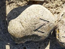 A large, weathered rock with a distinct carved symbol resembling the letter 'Z' on its surface, surrounded by sandy ground. photo